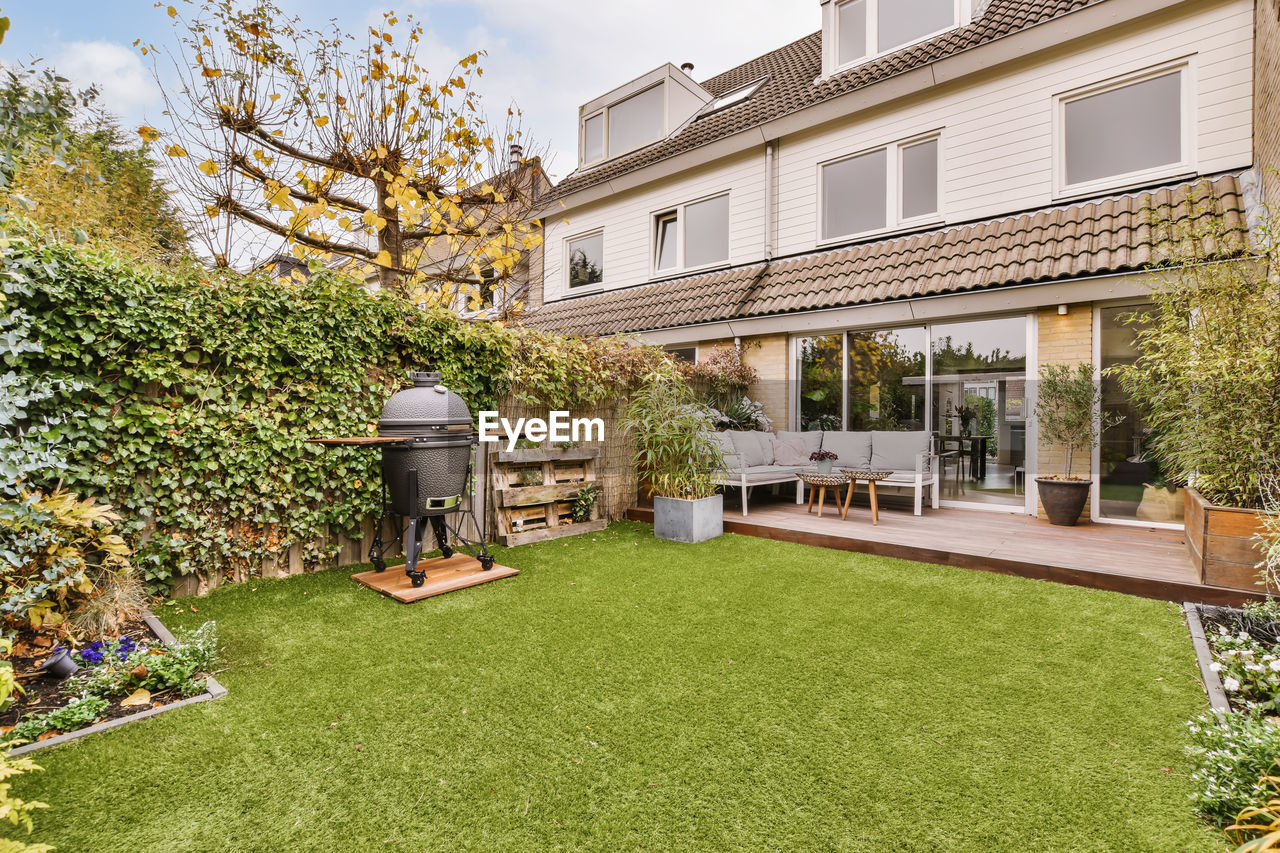 Cozy wooden table and soft sofa and grill placed in backyard with green garden near contemporary residential building