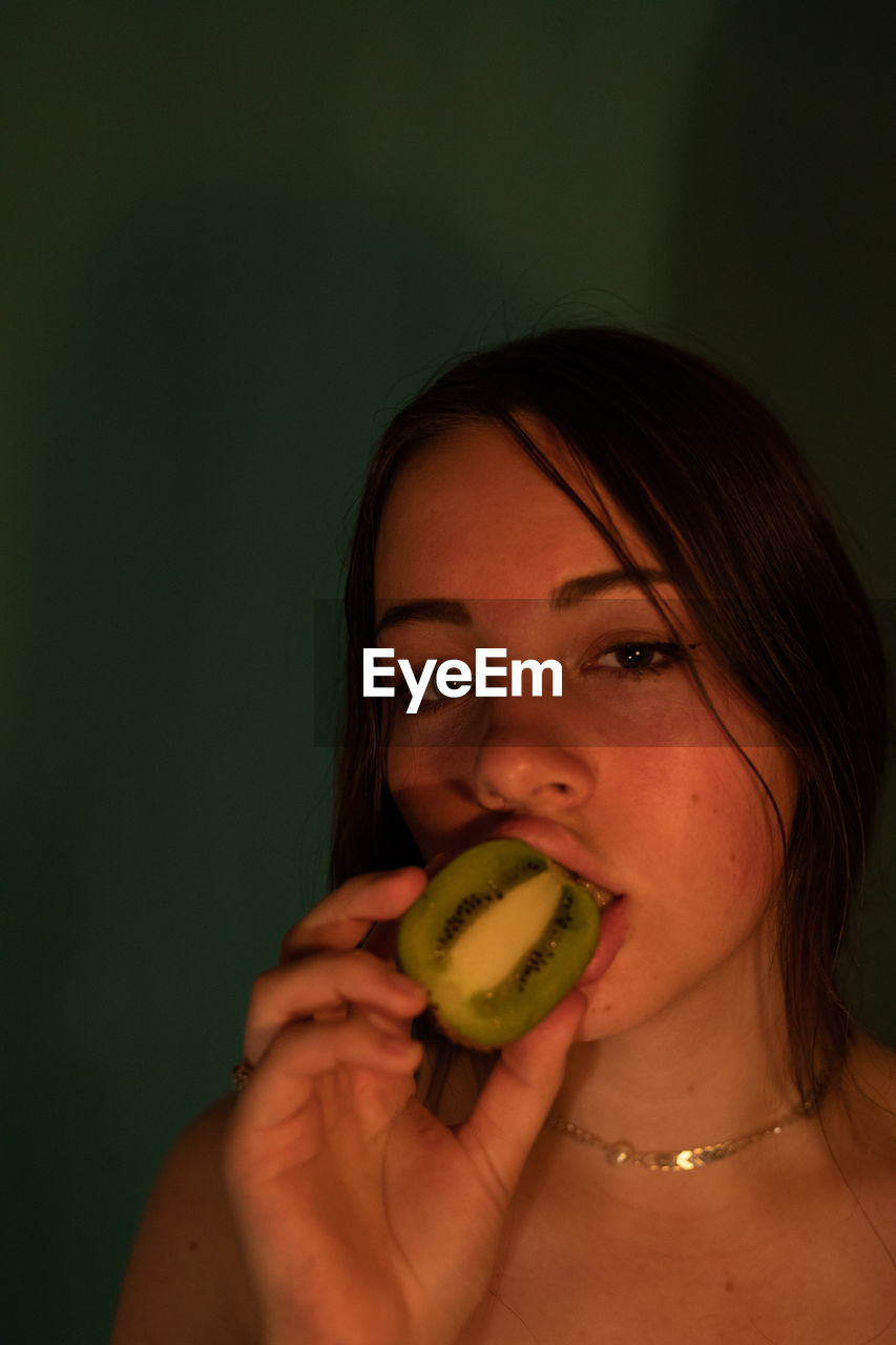 Portrait of woman eating kiwi against wall