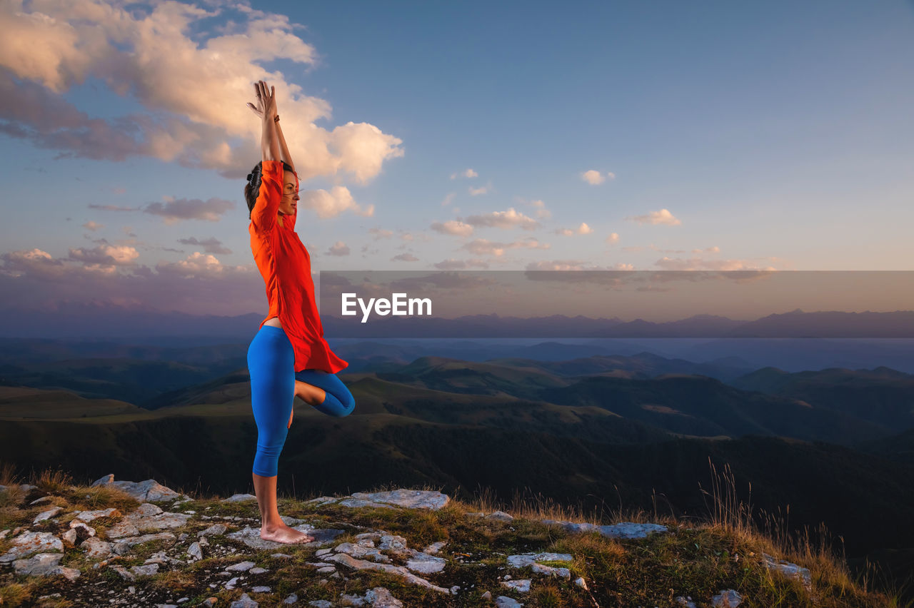Athletic woman stands in tree pose in yoga in the sunset light of the sun in the mountains