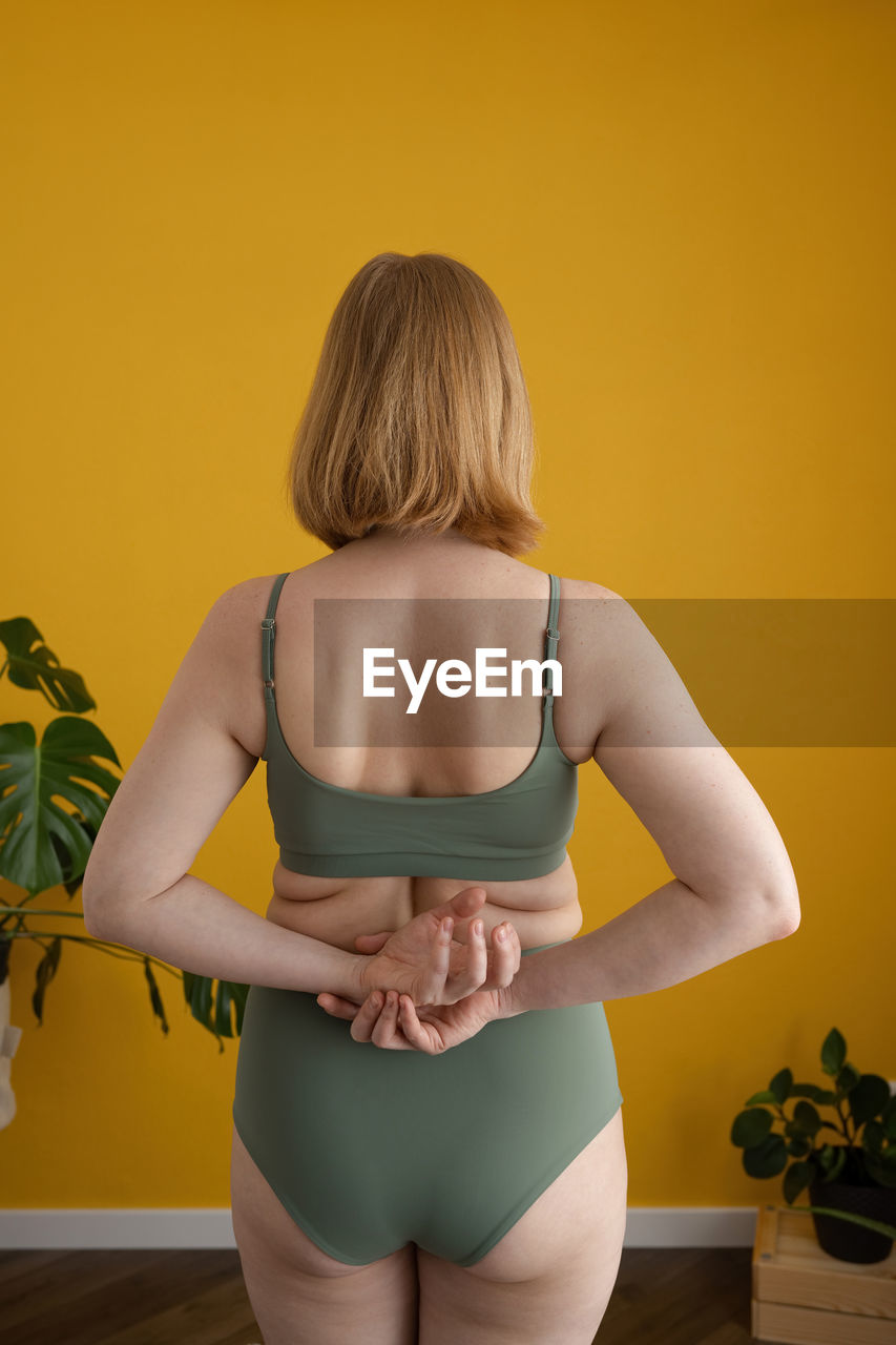 Back view of unrecognizable female with ginger hair and curvy body in lingerie standing against yellow wall at home