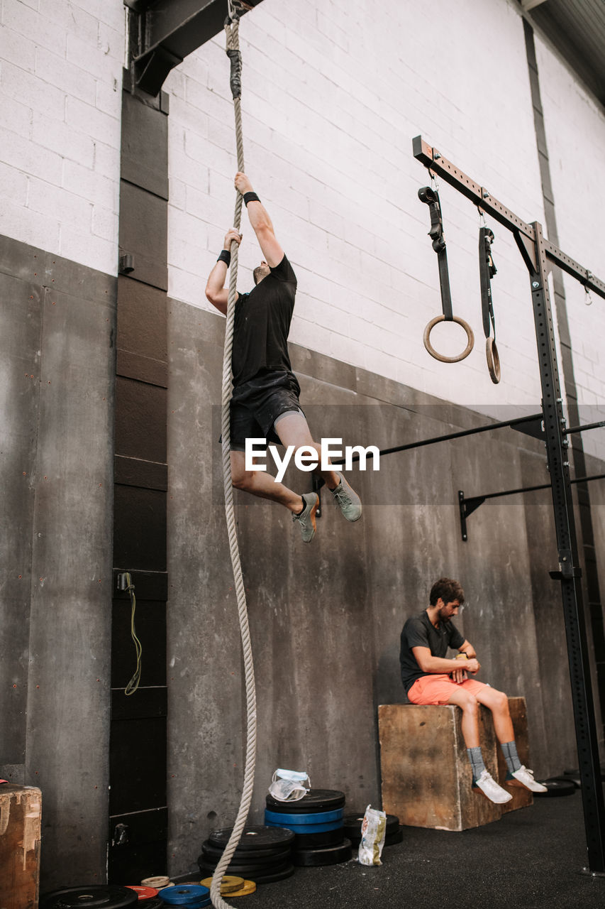 Low angle of muscular sportsman training in gym and climbing rope during functional workout