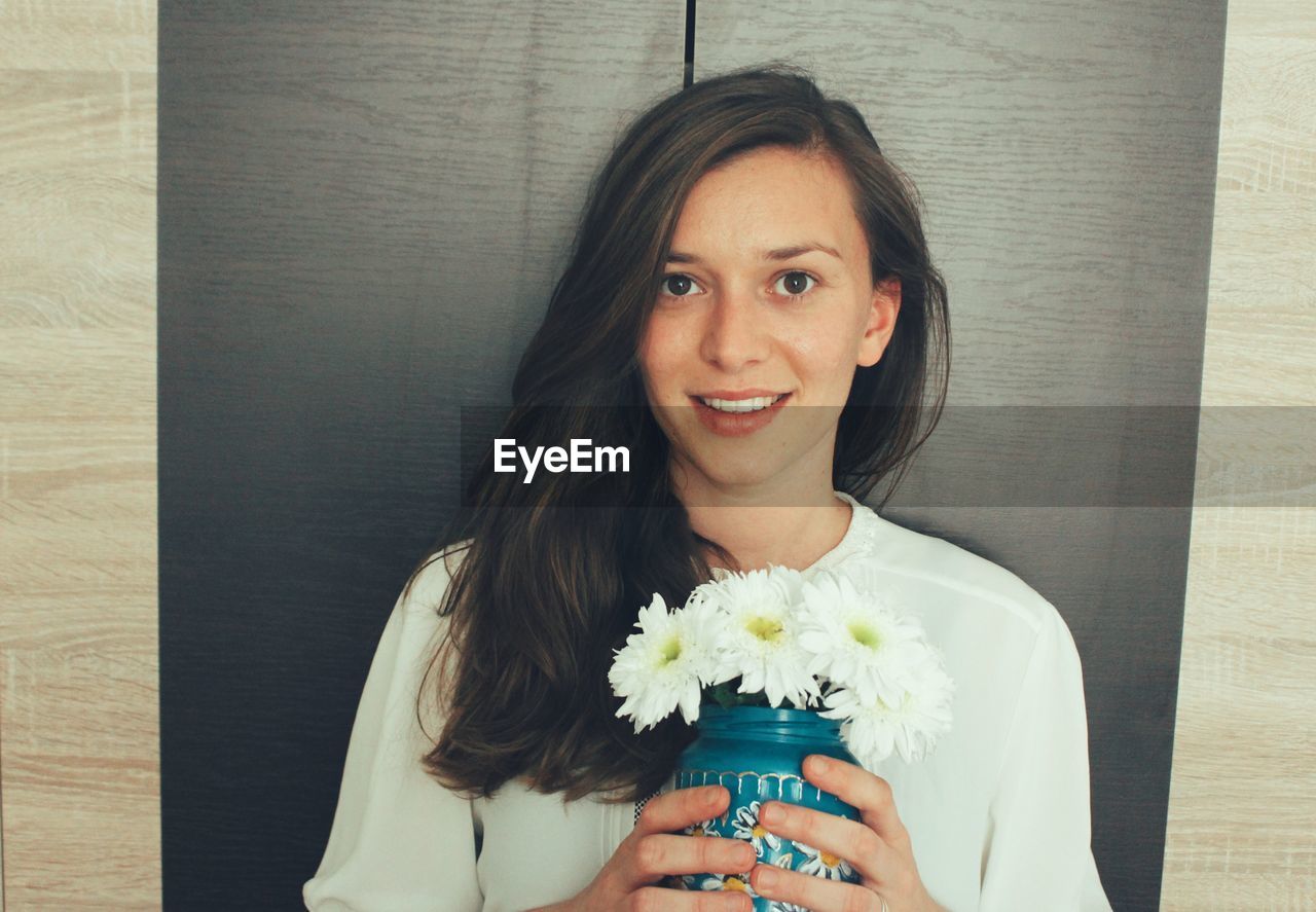 Portrait of smiling young woman holding flowers in vase at home