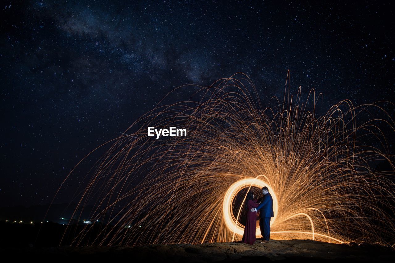 Full length side view of couple standing face to face by spinning wire wool against star field at night