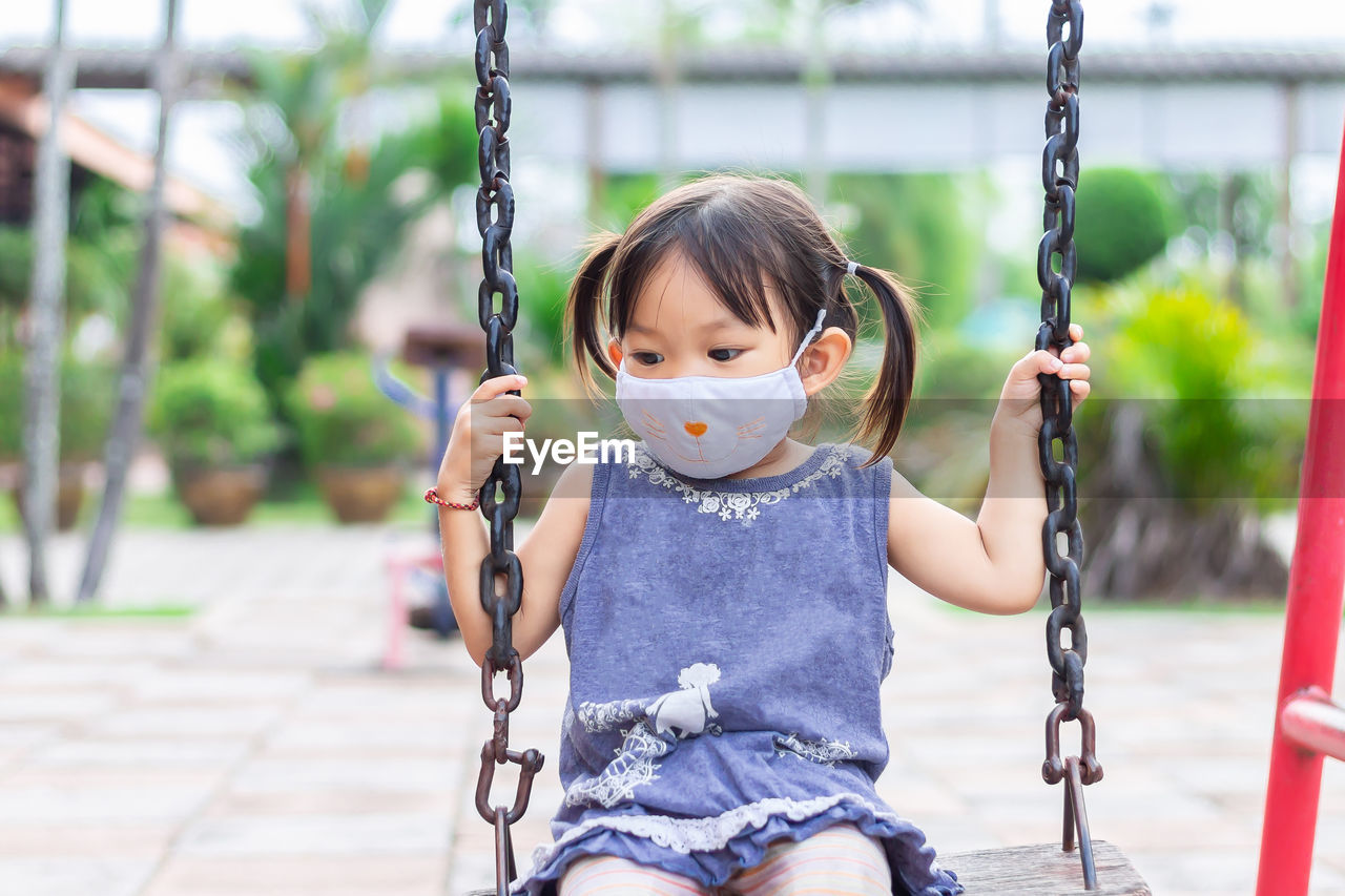 Girl wearing mask sitting on swing at playground