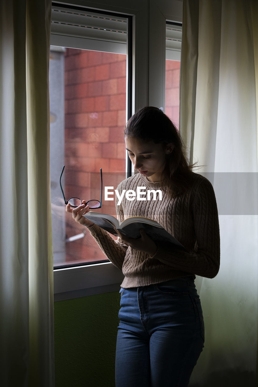 Young woman reading a book next to her window in the morning.