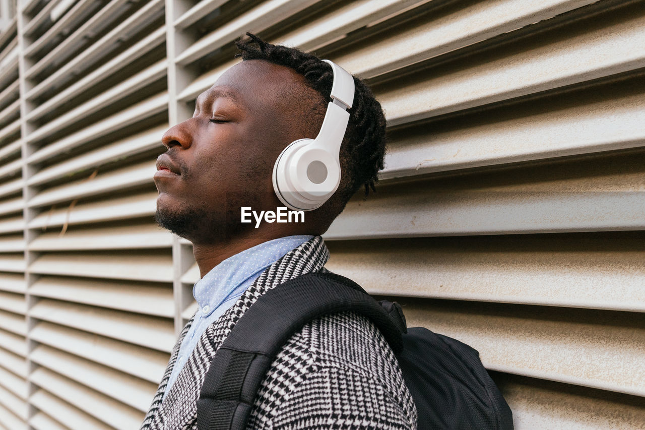 Side view of young mindful african american male office worker listening to song with closed eyes in wireless headset near ribbed wall in town