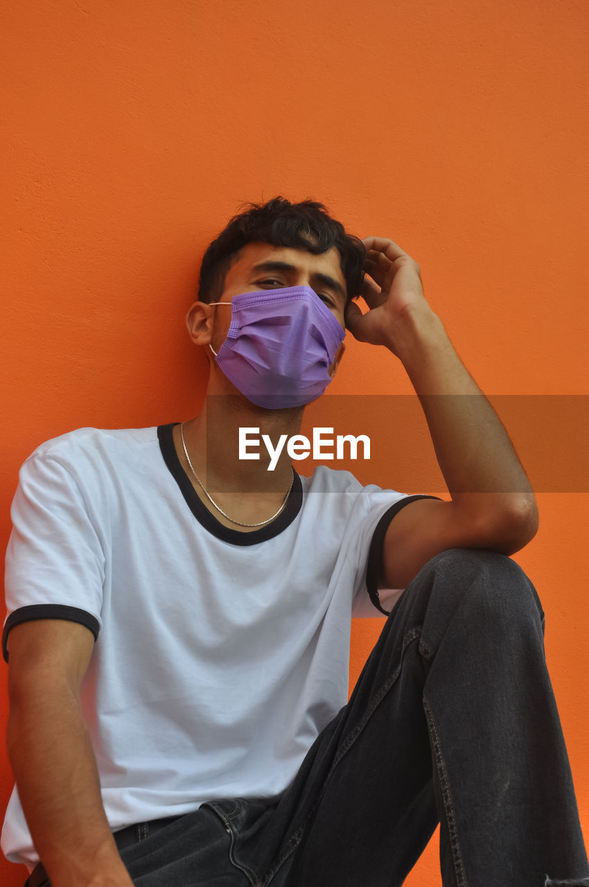 A indian young guy wearing face mask, looking at camera sitting against orange wall with copy space 