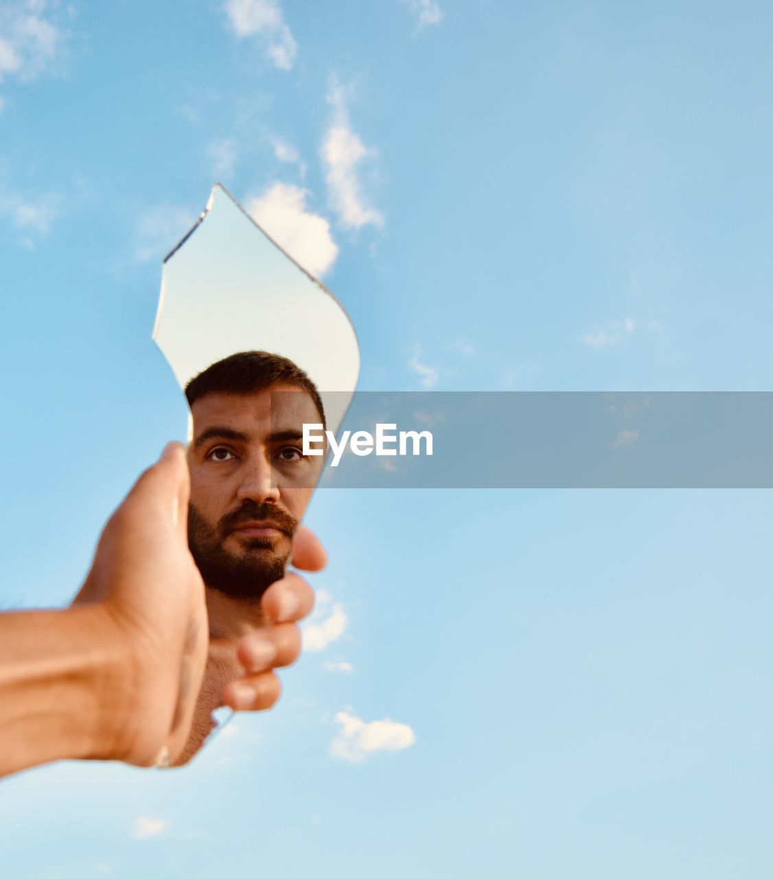 Cropped hand of man holding broken mirror against sky