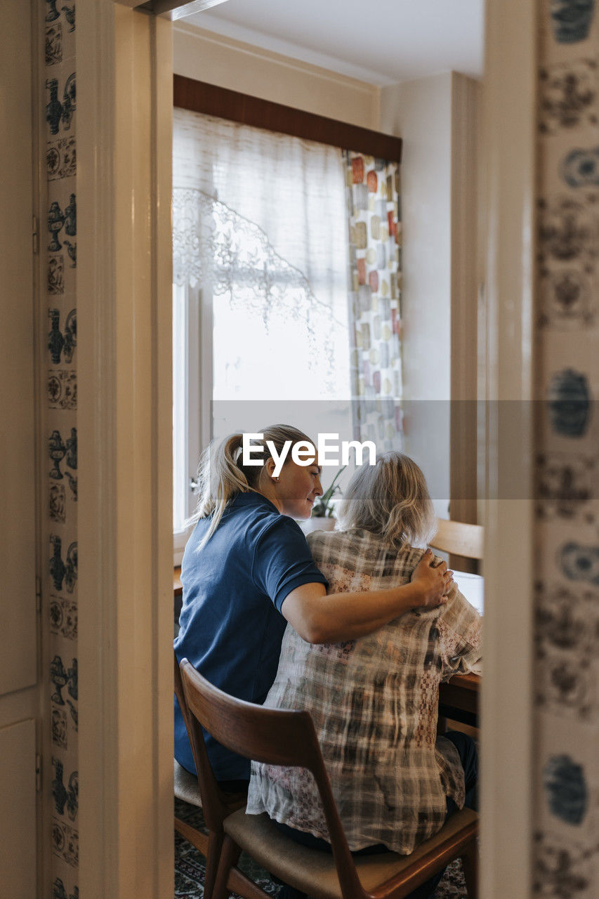 Female caregiver with arm around of senior woman sitting on chair at home