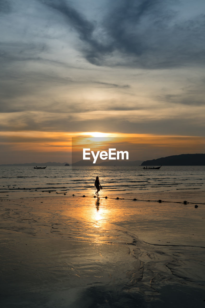 Silhouette person walking on shore at beach against sky during sunset