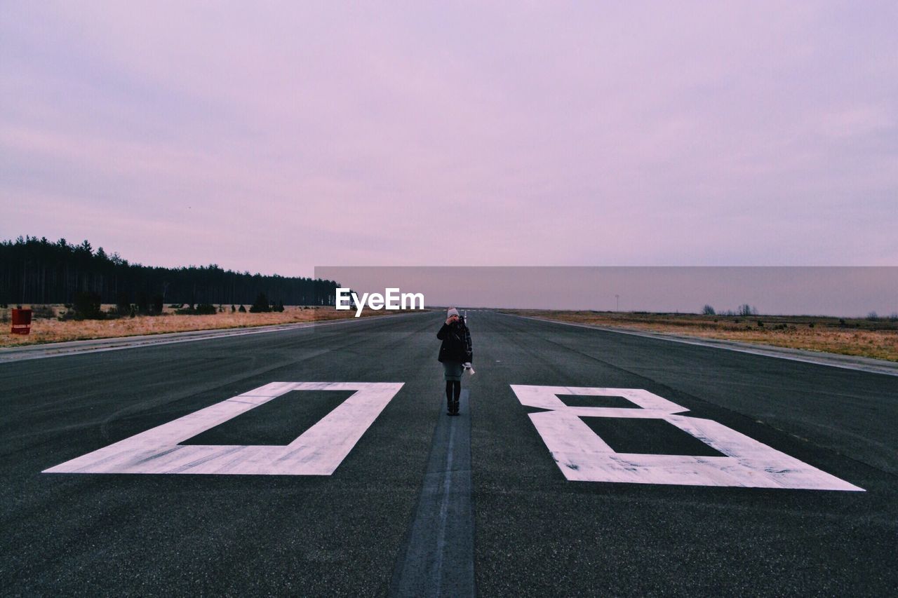 Man walking on airport runway against sky at sunset