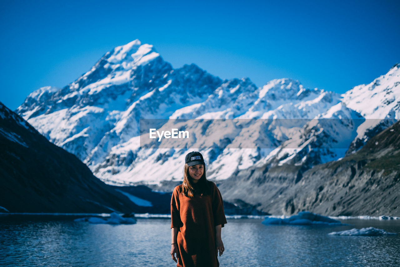 Young girl standing next lake shore with snowcapped mountain at the back.