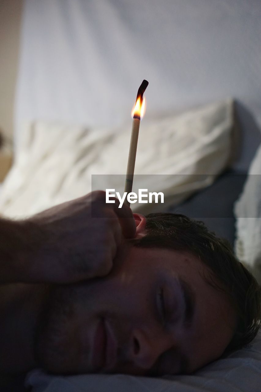 Cropped hand holding burning stick by ear of man lying on bed at home