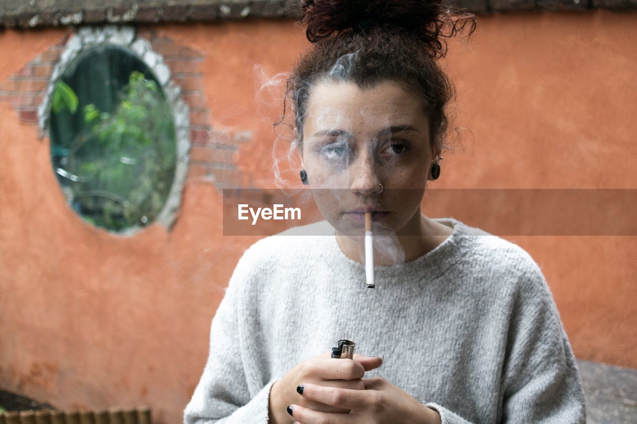 Young woman smoking while standing against wall