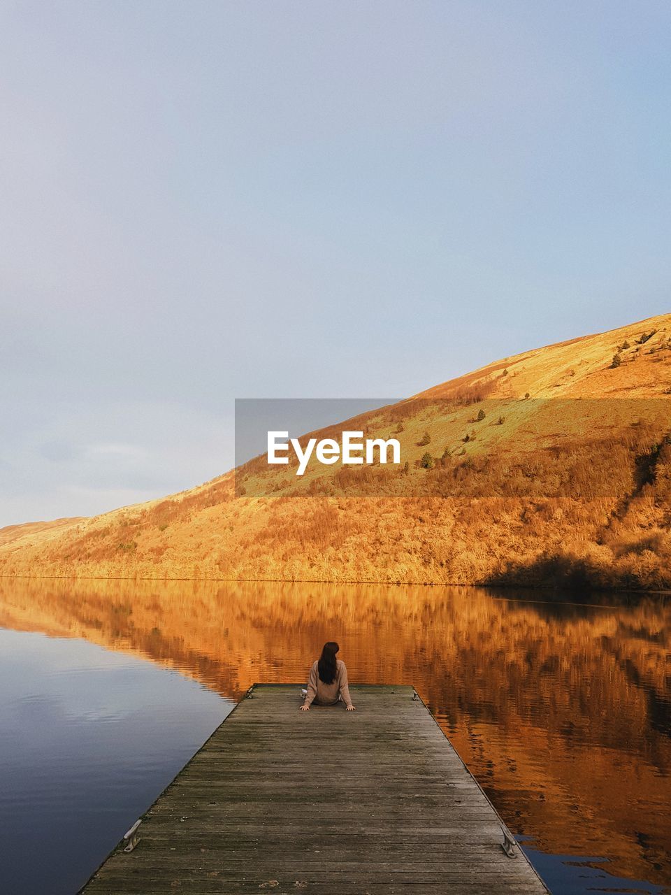 Rear view of woman sitting on pier over lake against sky