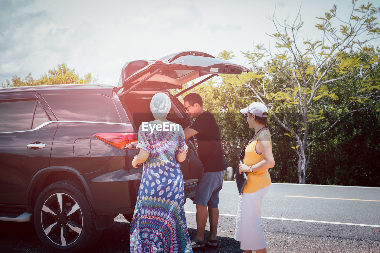 Man with female friends loading luggage in trunk of automobile on summer vacation trip