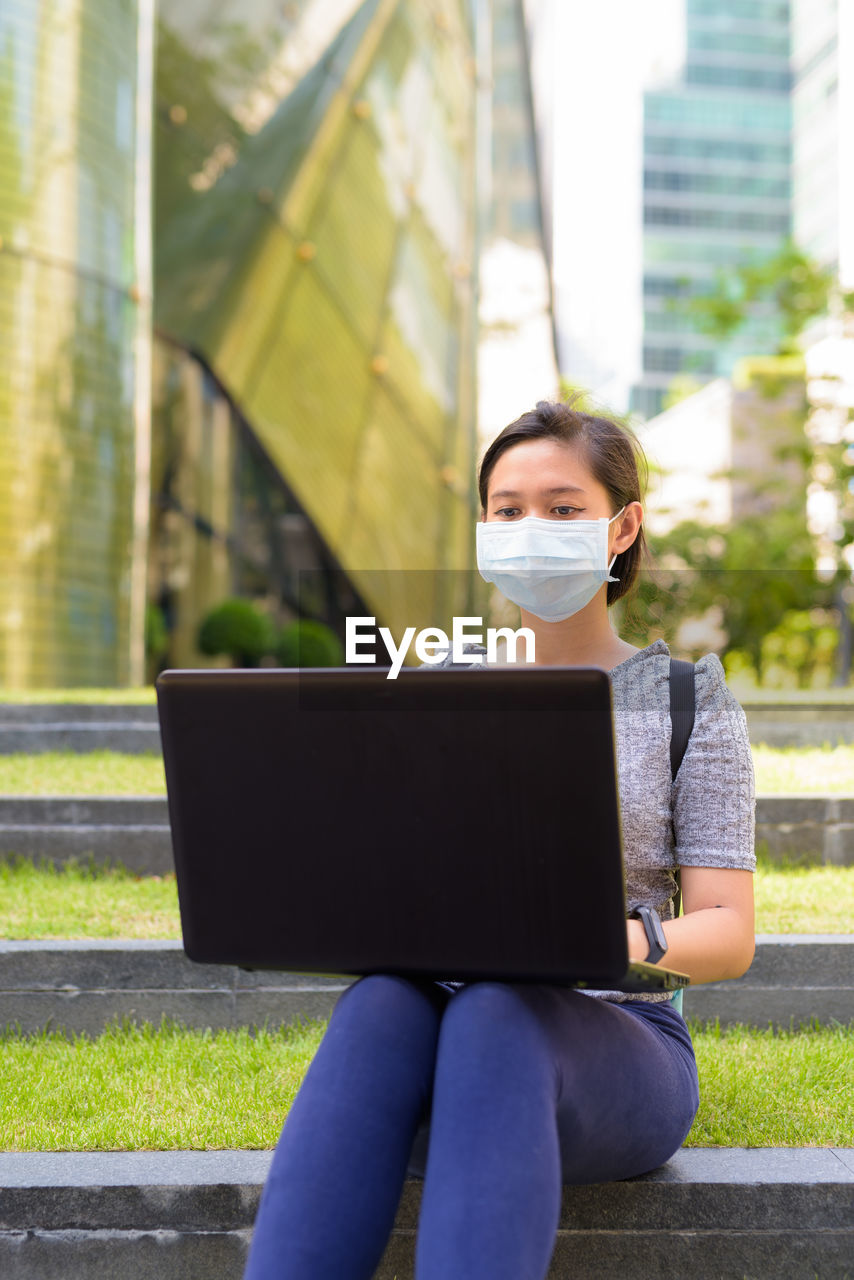 YOUNG WOMAN USING MOBILE PHONE WHILE SITTING ON LAPTOP OUTDOORS