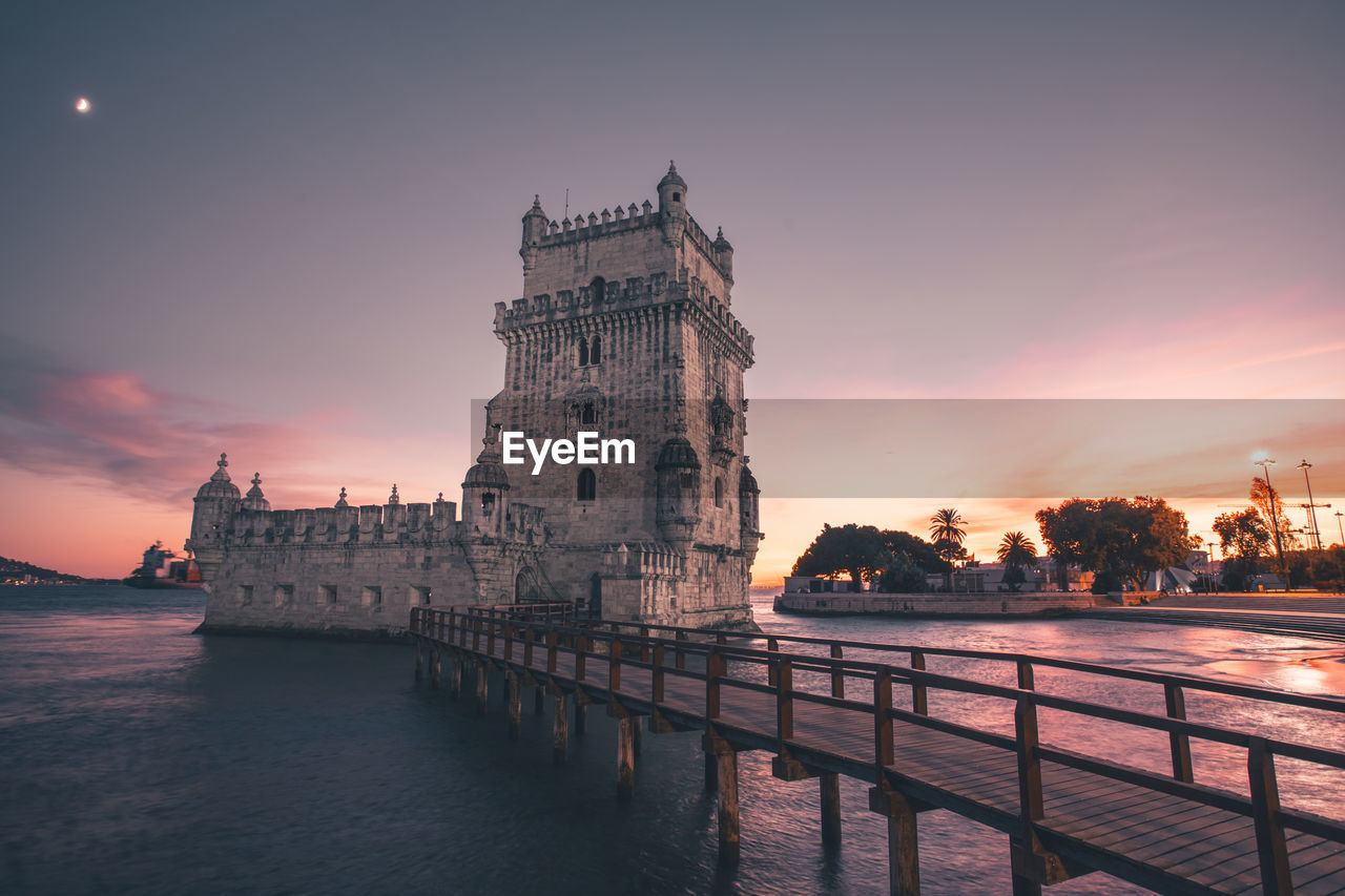 View of building by river against sky during sunset