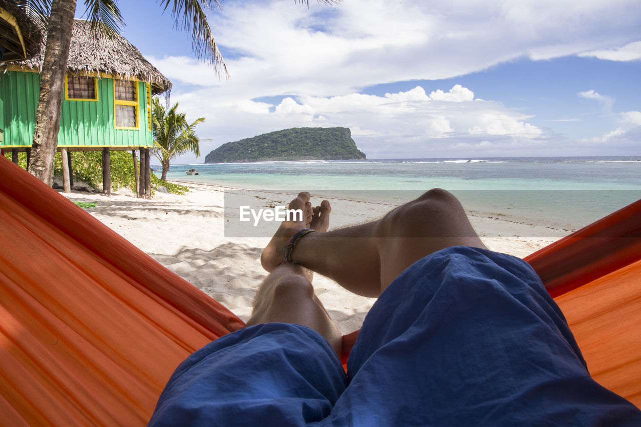 Pair of legs resting on orange hammock at white sandy beach