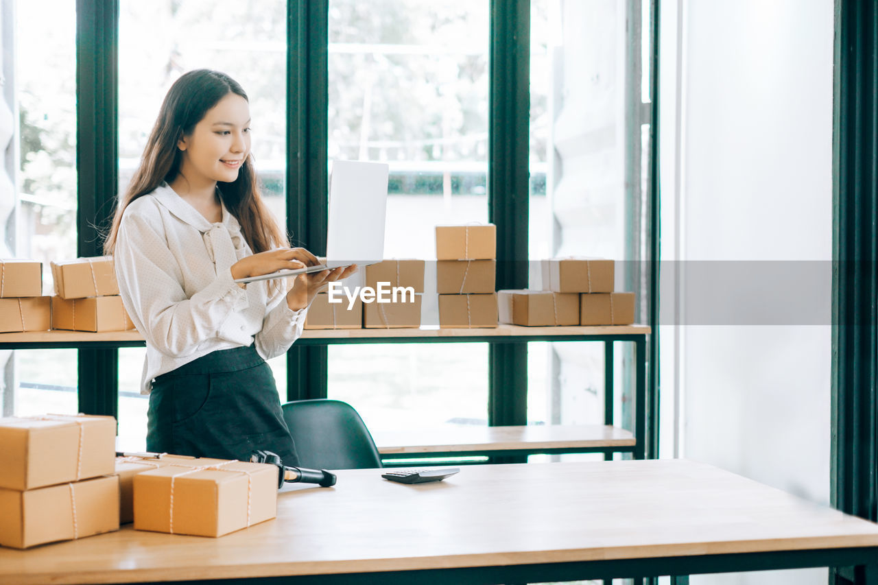 Young businesswoman using laptop by packages at office