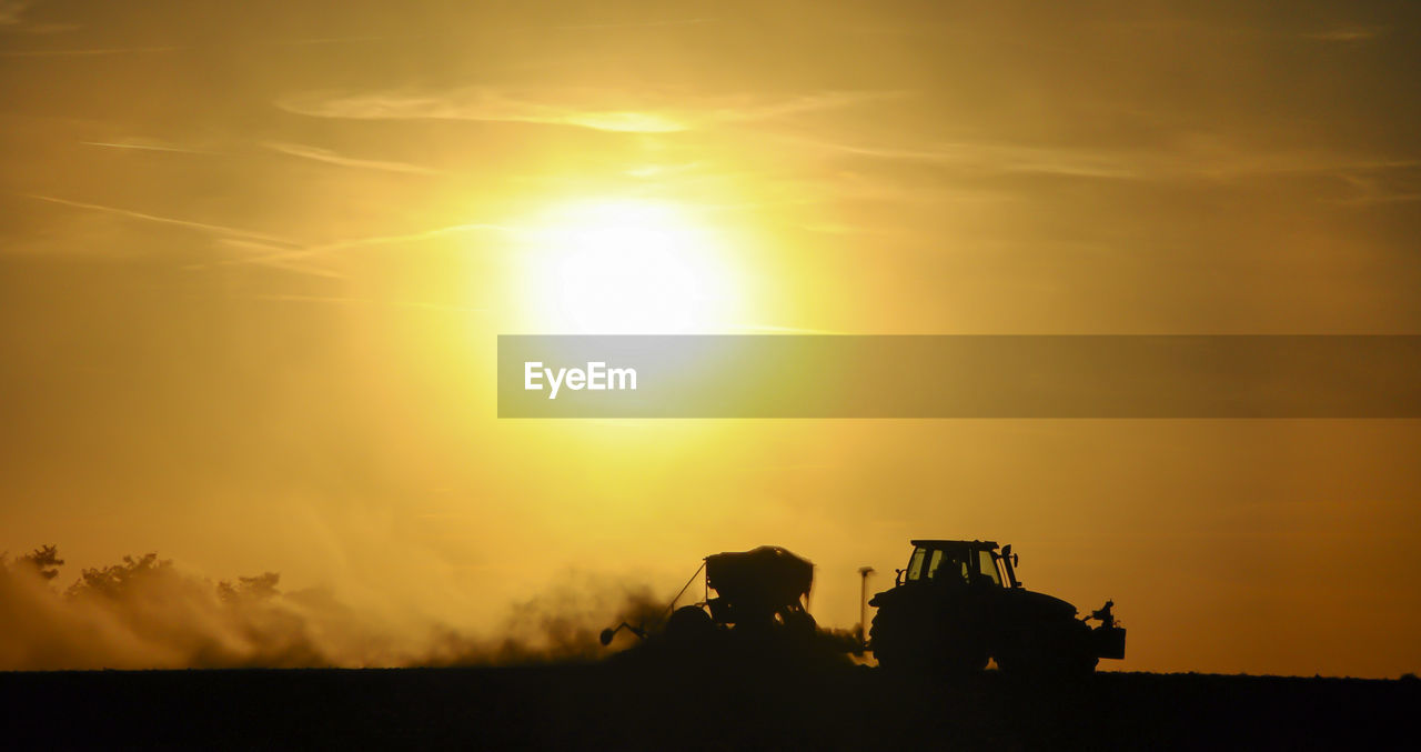 Silhouette of a tractor sowing seeds in a field in a cloud of dust against the background.