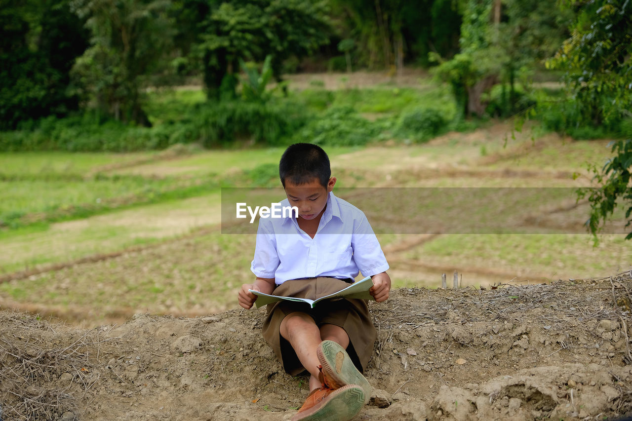 Full length of cute boy reading book while sitting on field against trees