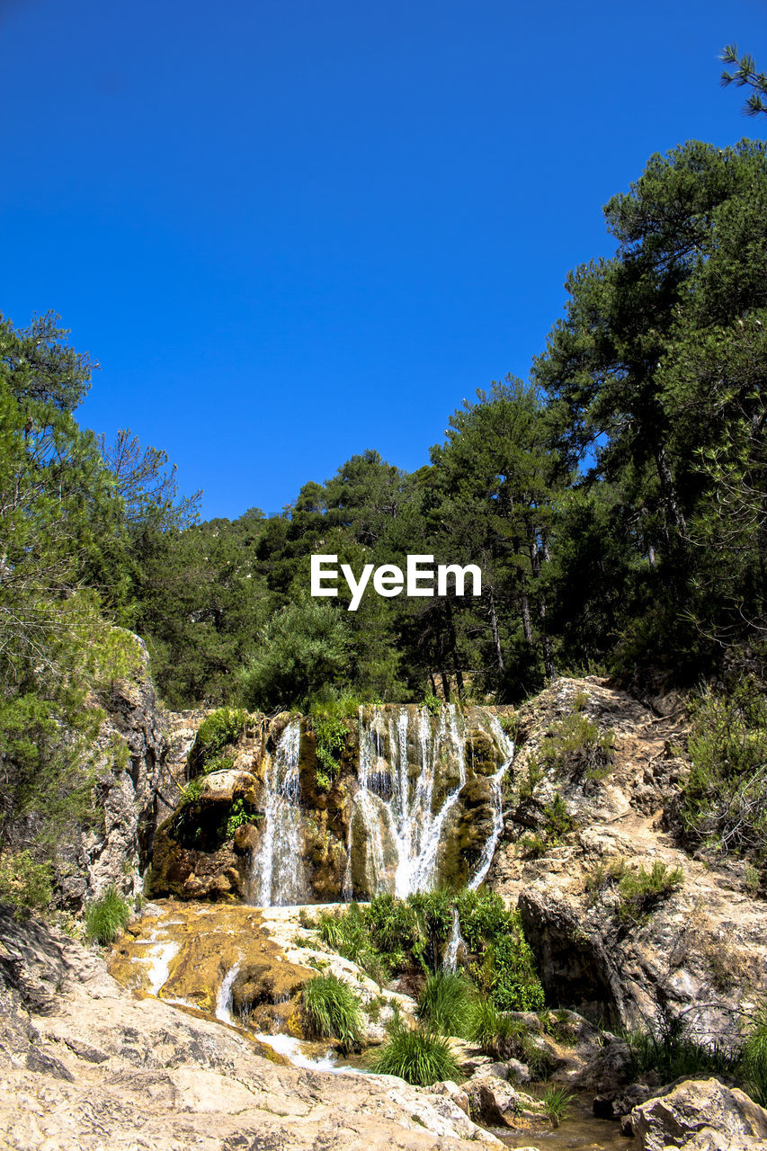 Scenic view of waterfall in forest against clear blue sky