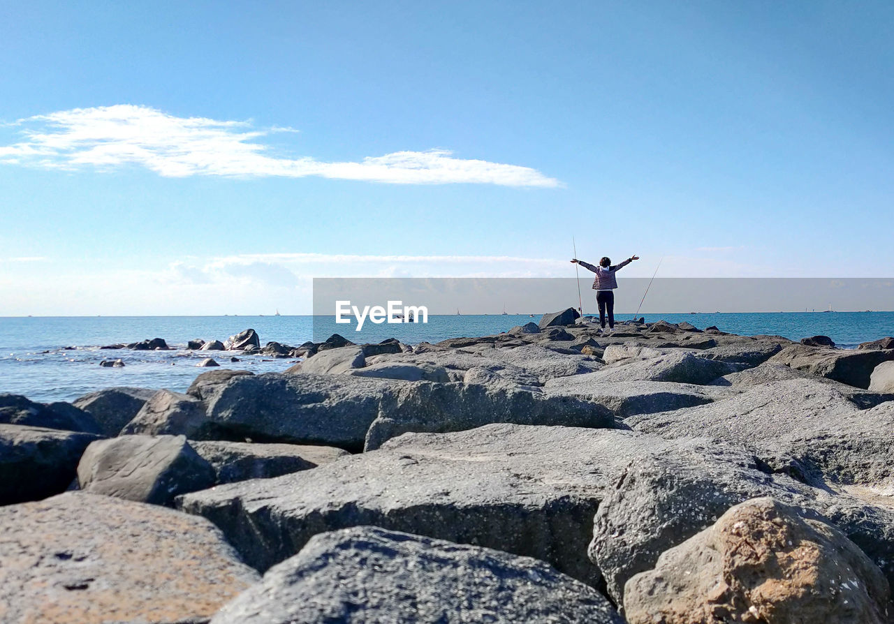 MAN STANDING ON ROCK AT BEACH AGAINST SKY