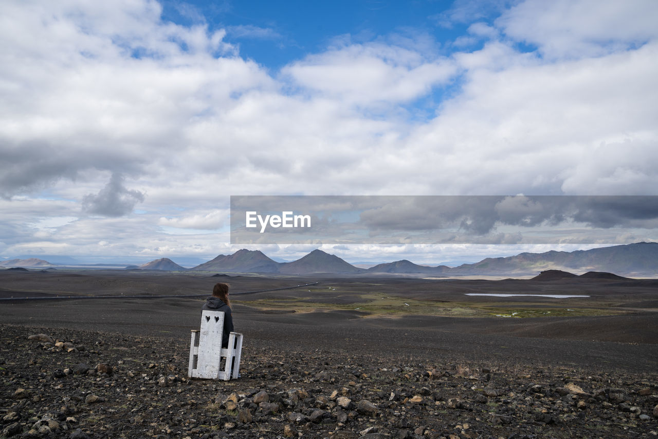 Rear view of woman sitting on chair at landscape against cloudy sky