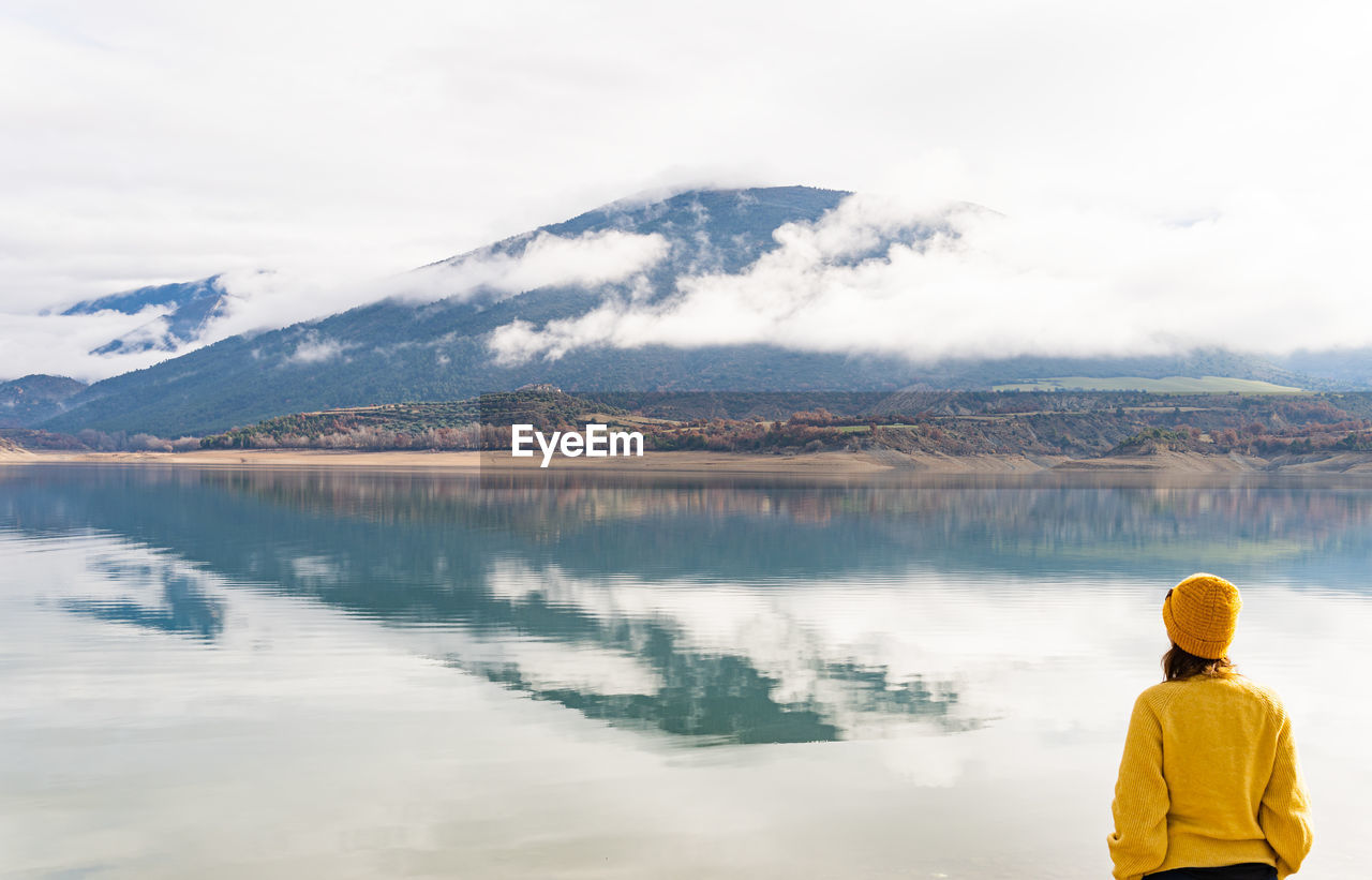 Rear view of woman wearing a sweater watching the mountains and cloud reflecting in a lake
