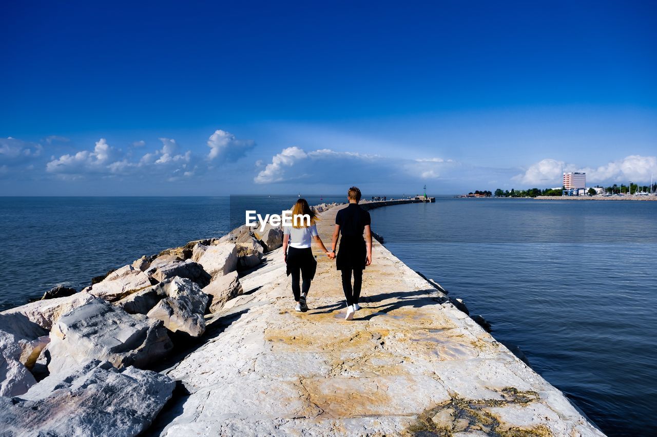 Rear view of couple holding hands walking on pier over sea