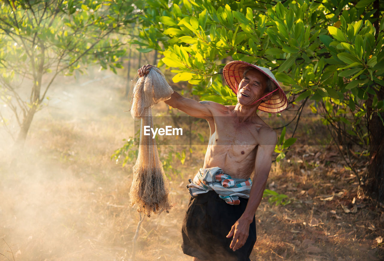 Shirtless senior man holding fishing net at forest