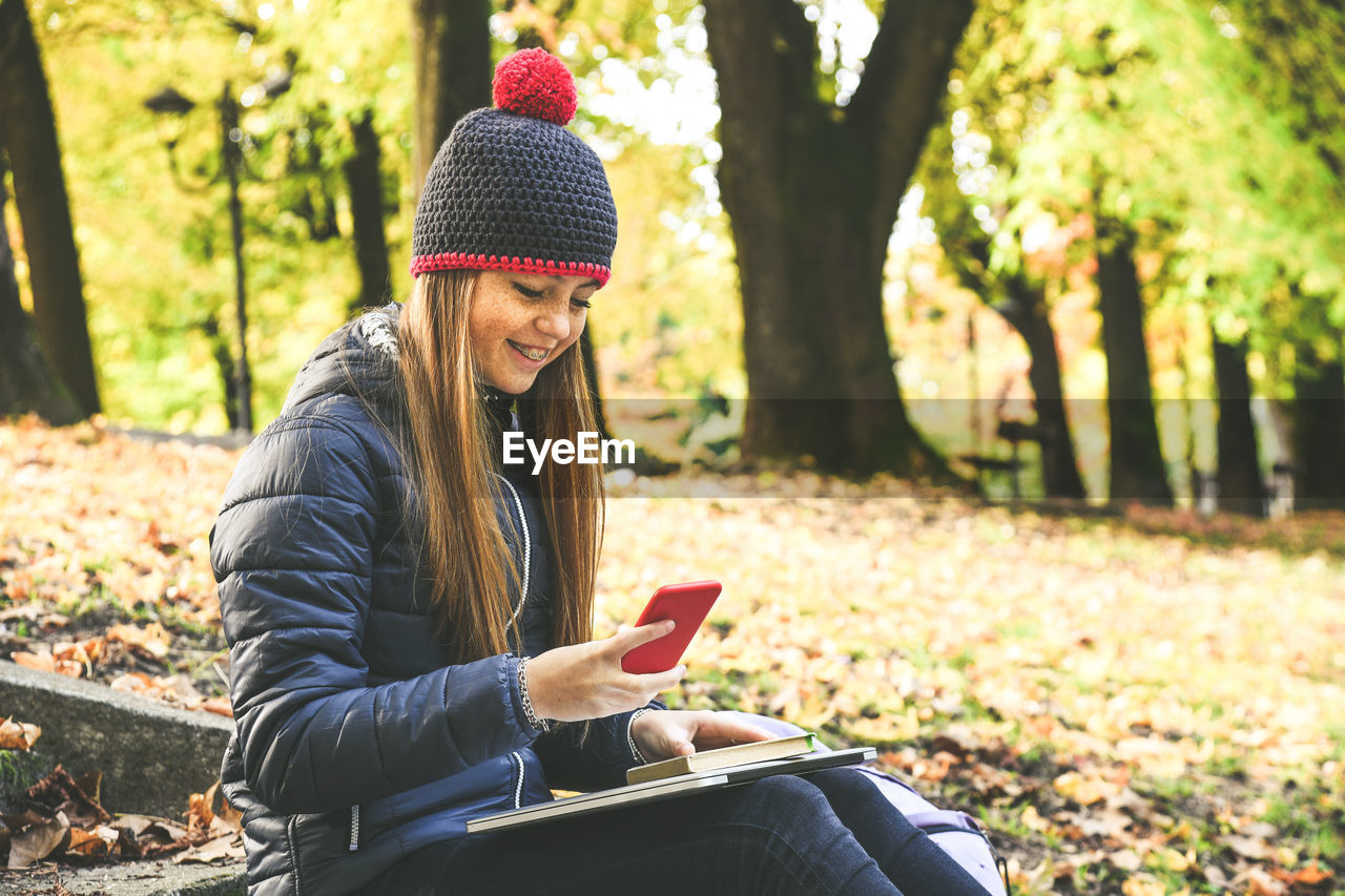 Girl with wool cap sitting in the park using smartphone. teen using mobile phone, chat with friends