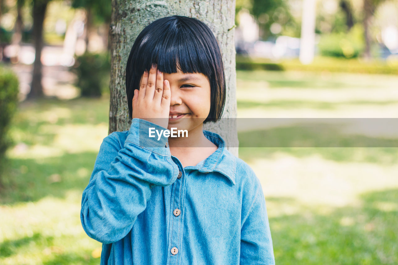 Portrait of smiling girl with hand covering eye against tree trunk