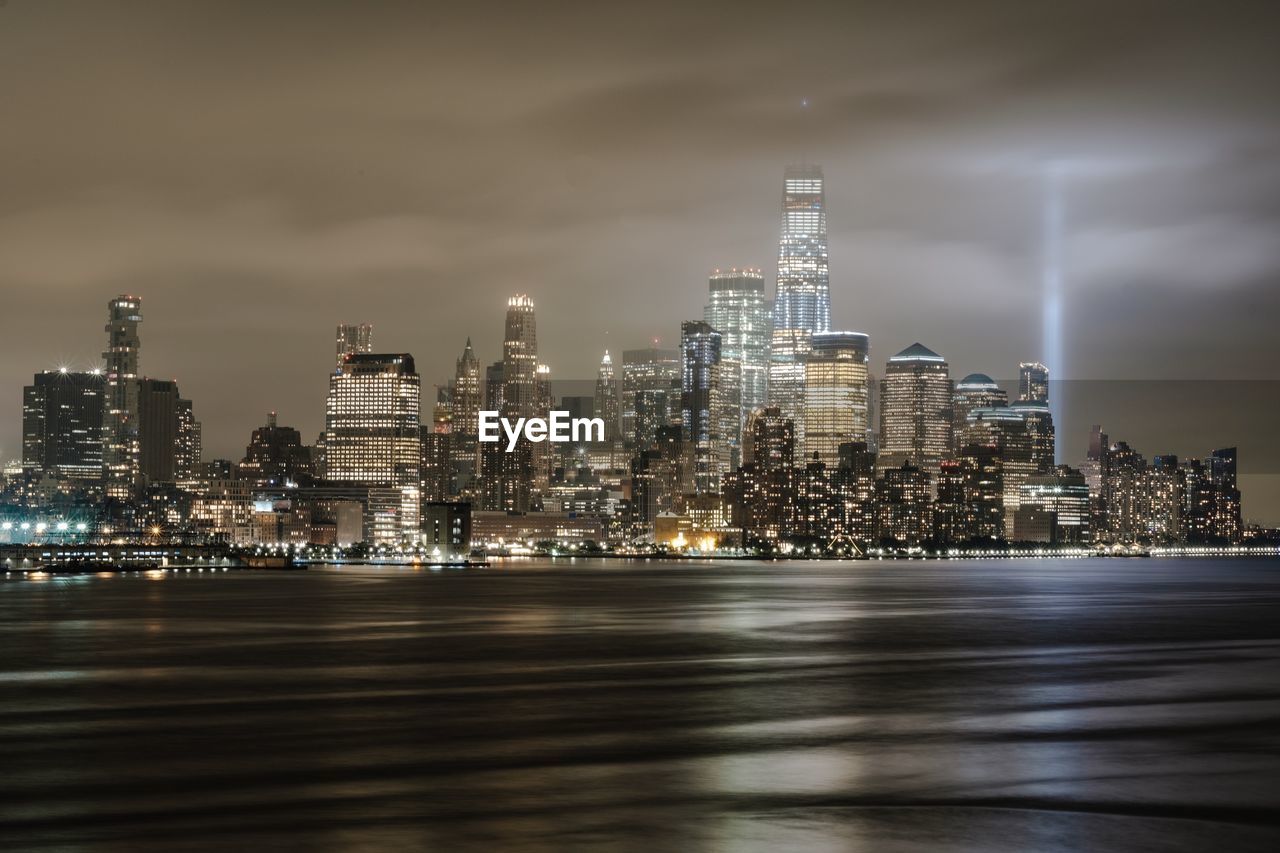 Illuminated city buildings by river against sky at night