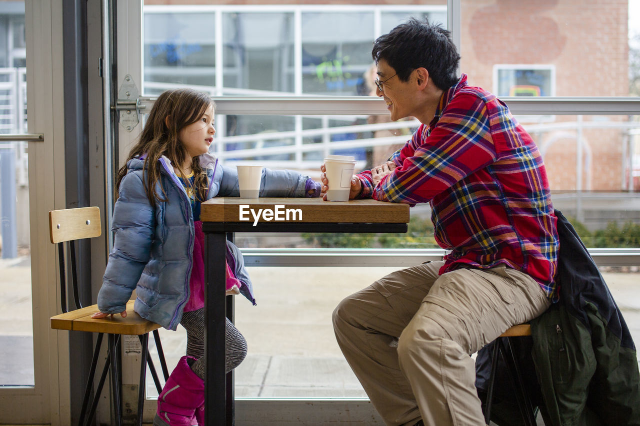 A little girl sits at a table by a large window with her happy father