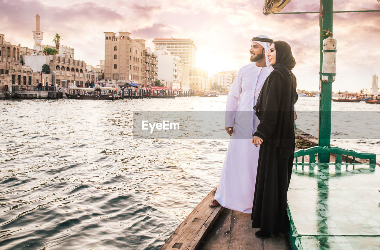 Smiling couple looking at view while standing in boat on river during sunset
