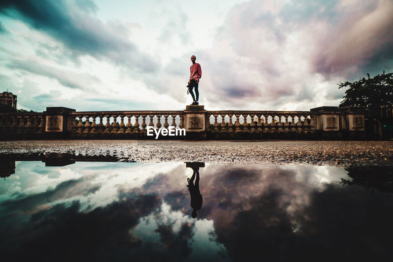 Low angle view of man standing on retaining wall with reflection on pond