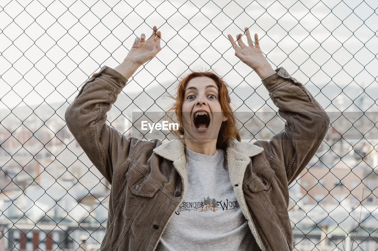 Young beautiful woman screaming looking at camera in front of metal fence with city view behind