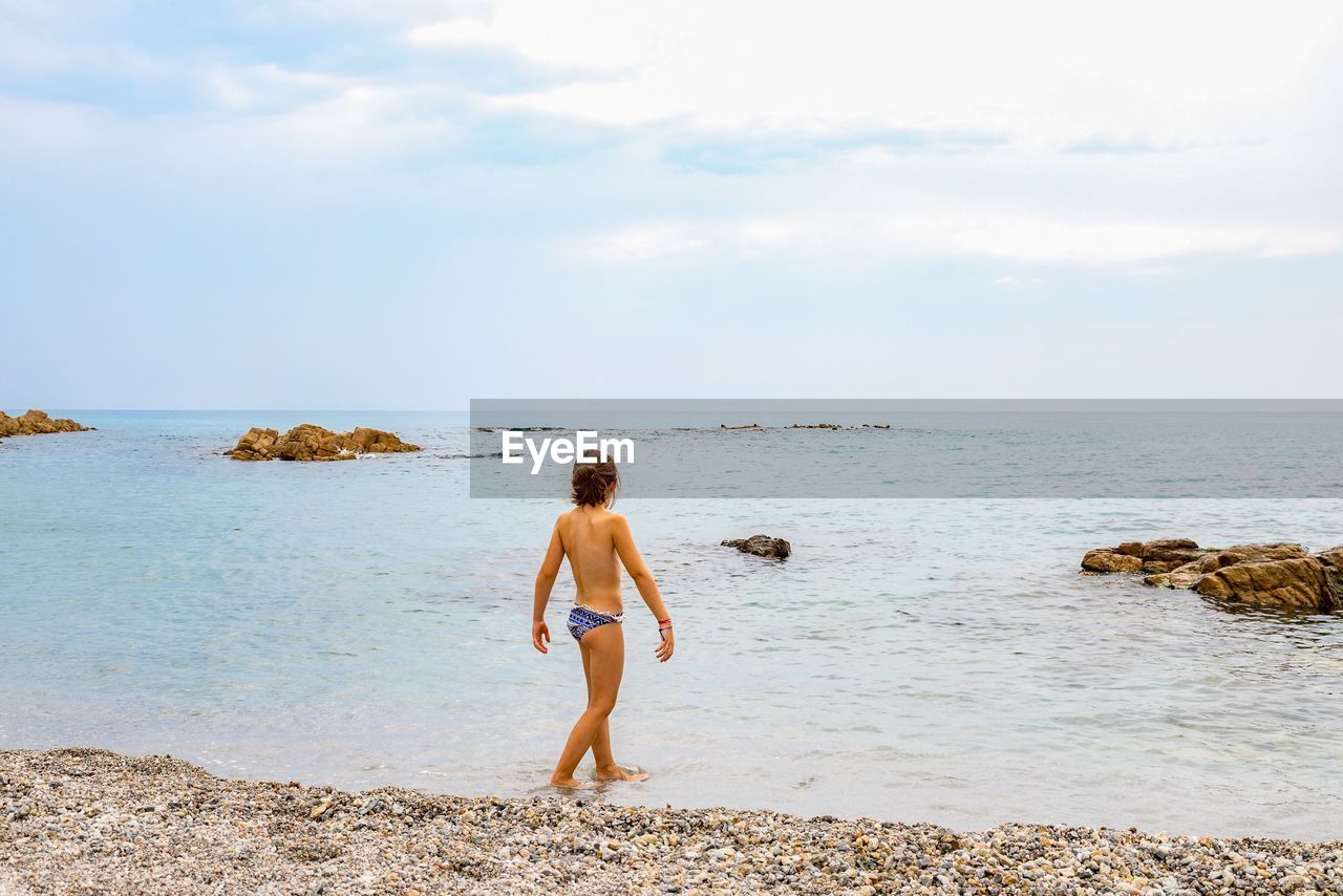 Shirtless woman standing on shore against sky