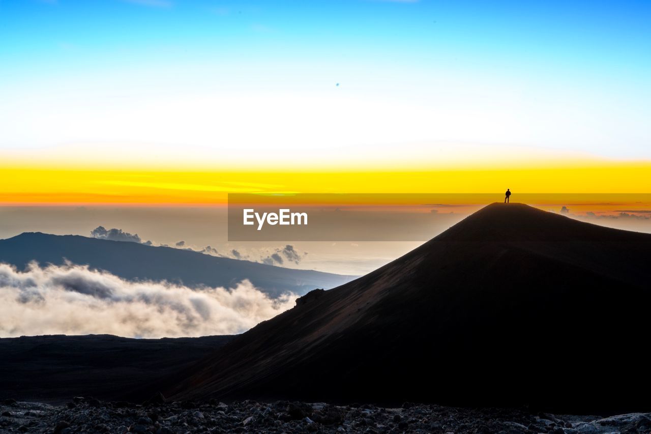 Distant view of man standing on mountain against sky during sunset