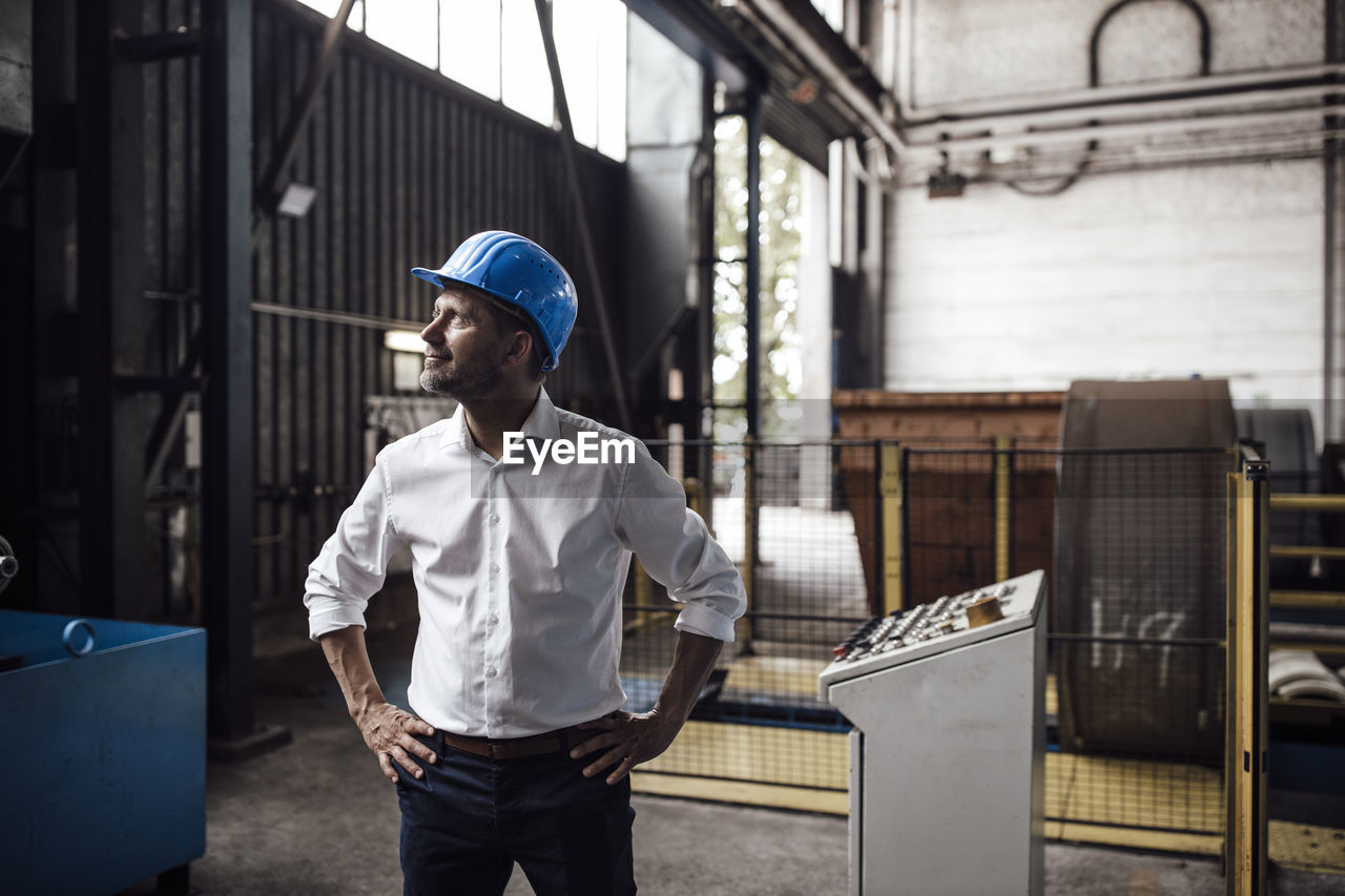 Smiling businessman with hands on hip looking up while standing in industry