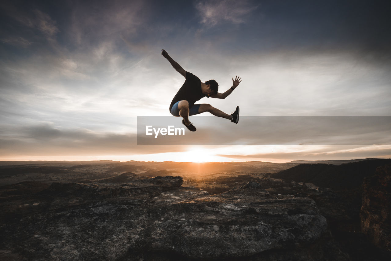 Full length of man jumping over mountain against cloudy sky at sunset