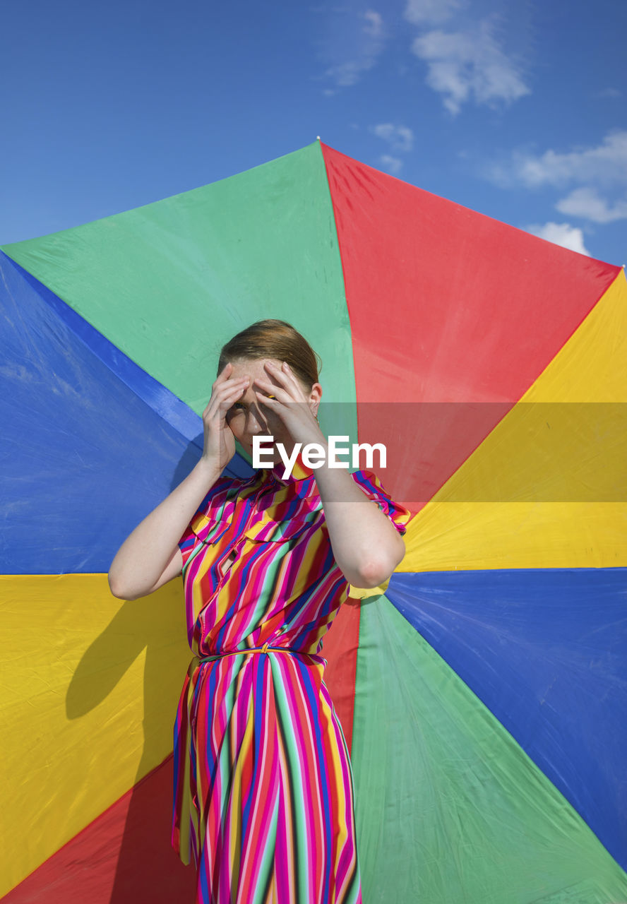 Woman shielding eyes while standing against colorful beach umbrella