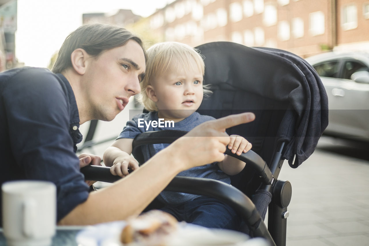 Father pointing while looking away with son at sidewalk cafe in city