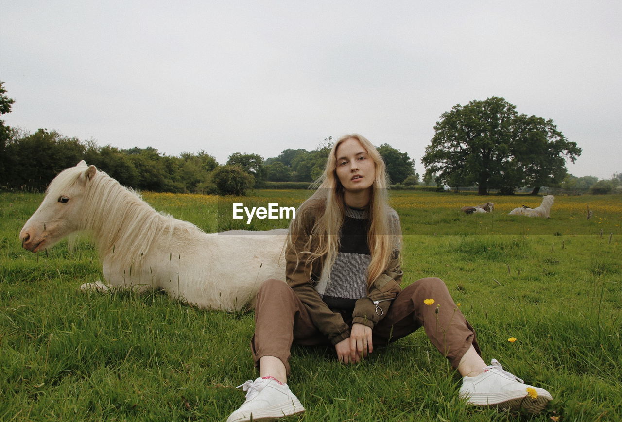 Portrait of woman sitting by horse on field against sky