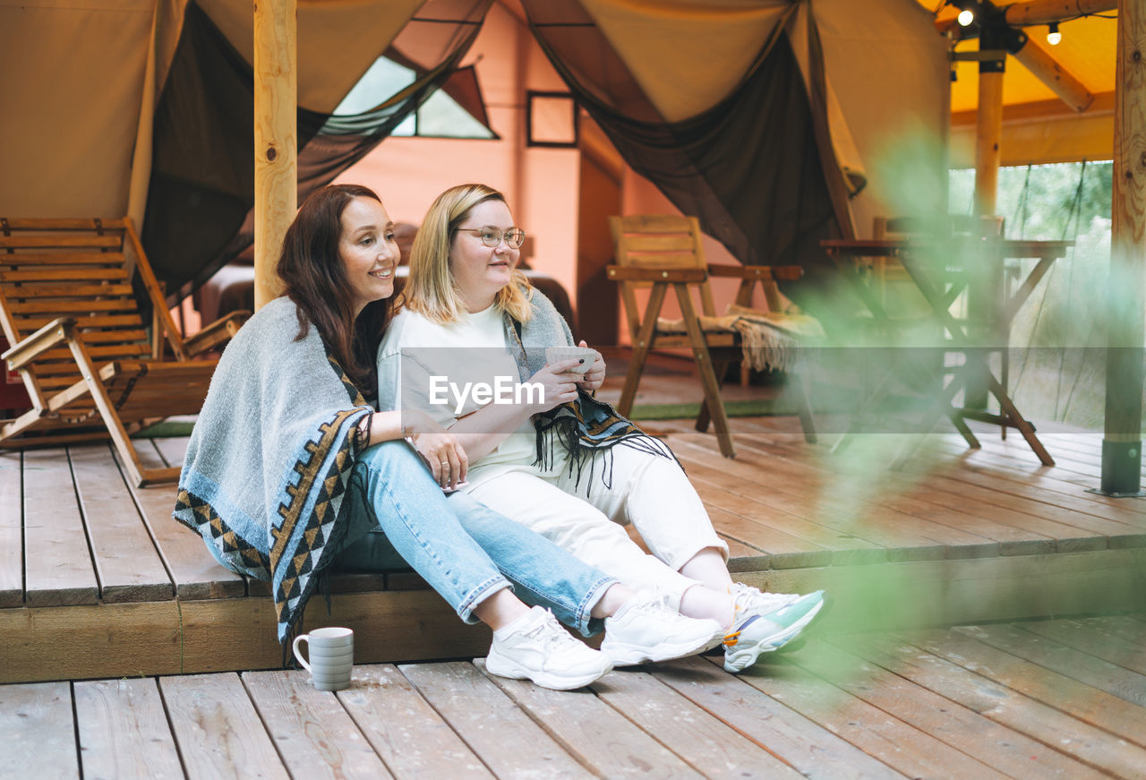 Two young women friends drinking tea and relaxing in glamping in the woods