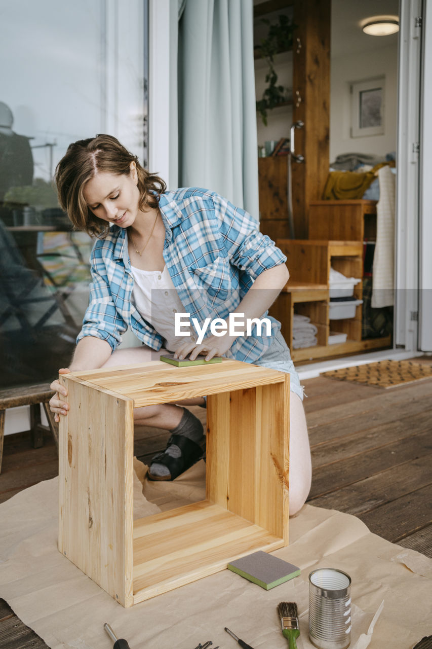 Young woman making wooden furniture while kneeling on porch outside house