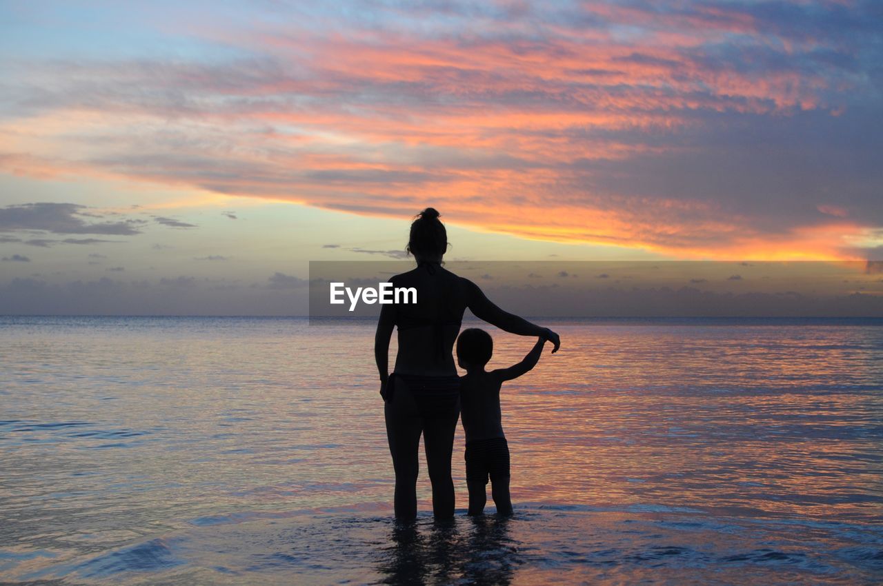 Rear view of silhouette of mother and son on beach against sunset sky