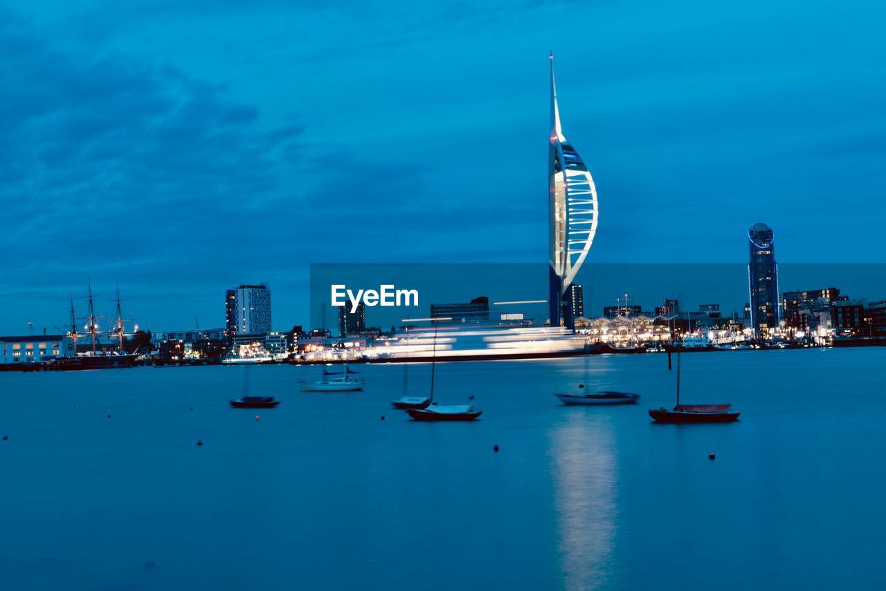 Sailboats in sea by modern buildings against blue sky